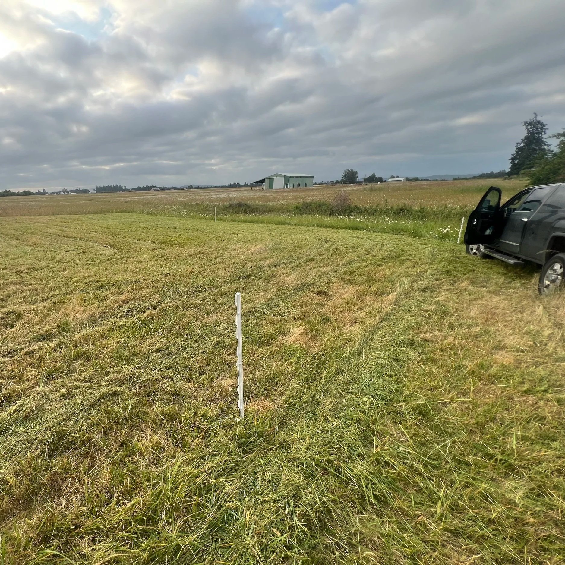 IMG_8943+(2) A grassy field with a freshly mowed area, a white marker in the foreground, and a black pickup truck parked nearby. In the background, a green building and overcast sky complete the rural landscape.