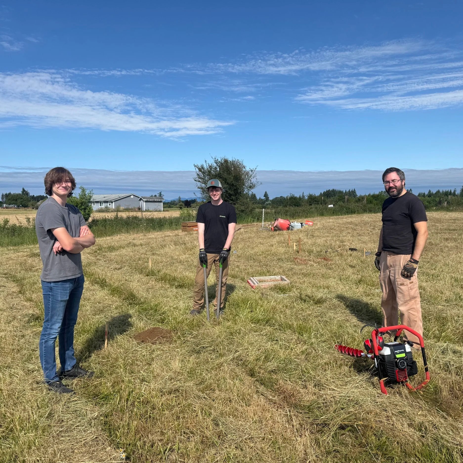 IMG_9518 Three individuals working on a grassy field, preparing for a project. One person operates a post hole digger, while the others stand nearby, ready to assist. In the background, tools and equipment are visible, along with a clear blue sky and distant trees.