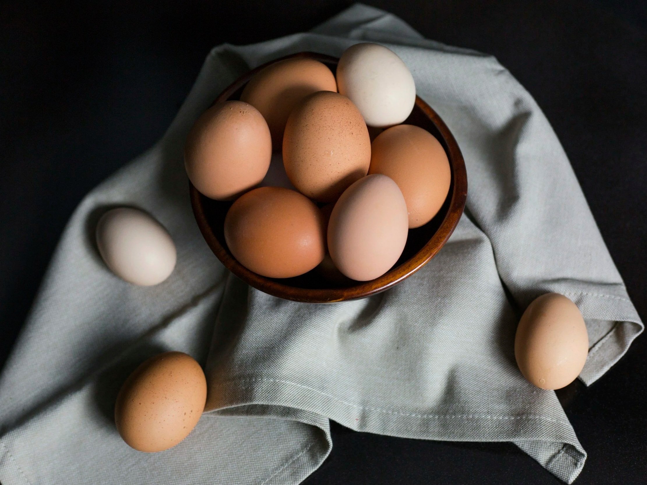 unsplash-image-BzF1XBy5xOc (2) A wooden bowl filled with various eggs, including brown and white, placed on a light green cloth against a dark background.